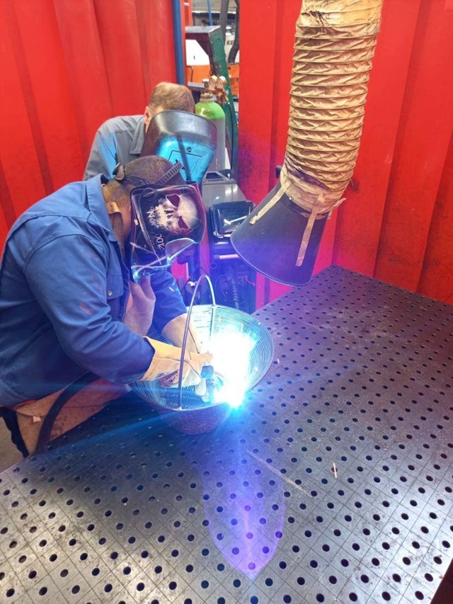 Zwei Personen führen Schweißarbeiten an einem Metallbauteil in einer Werkstatt durch; ein heller Lichtbogen entsteht, während eine Absauganlage und rote Schutzvorhänge die Umgebung prägen.