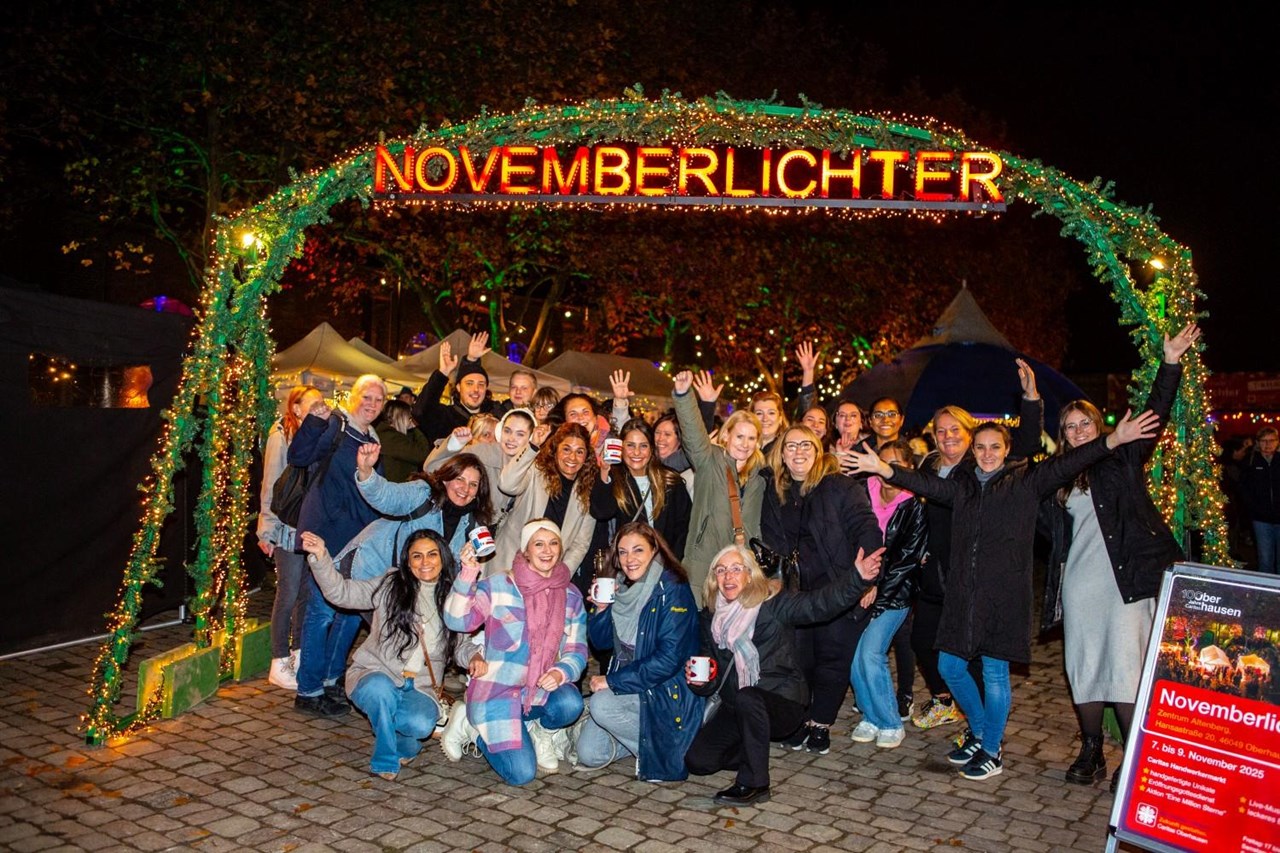 Eine große Gruppe Menschen, vor allem Frauen, steht unter einem mit künstlichem Tannengrün und Lichterketten dekorierten Holzbogen, an dem der leuchtend rote Schriftzug "Novemberlichter" angebracht ist.