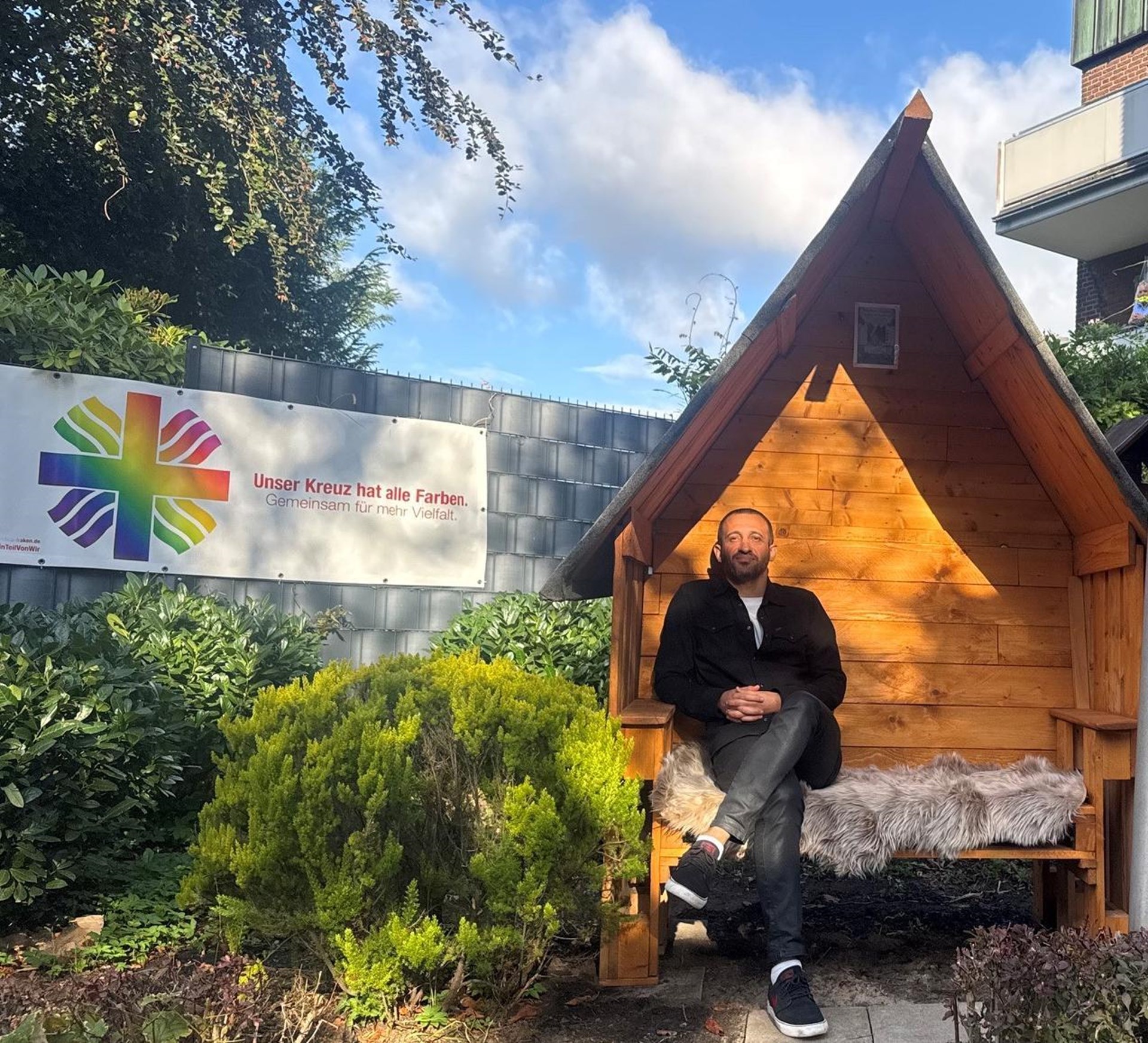 Person sitzt in einem hölzernen Wartehäuschen in einem Garten. Daneben ein Schild mit regenbogenfarbenem Kreuz und dem Slogan „Unser Kreuz hat alle Farben. Gemeinsam für mehr Vielfalt.“