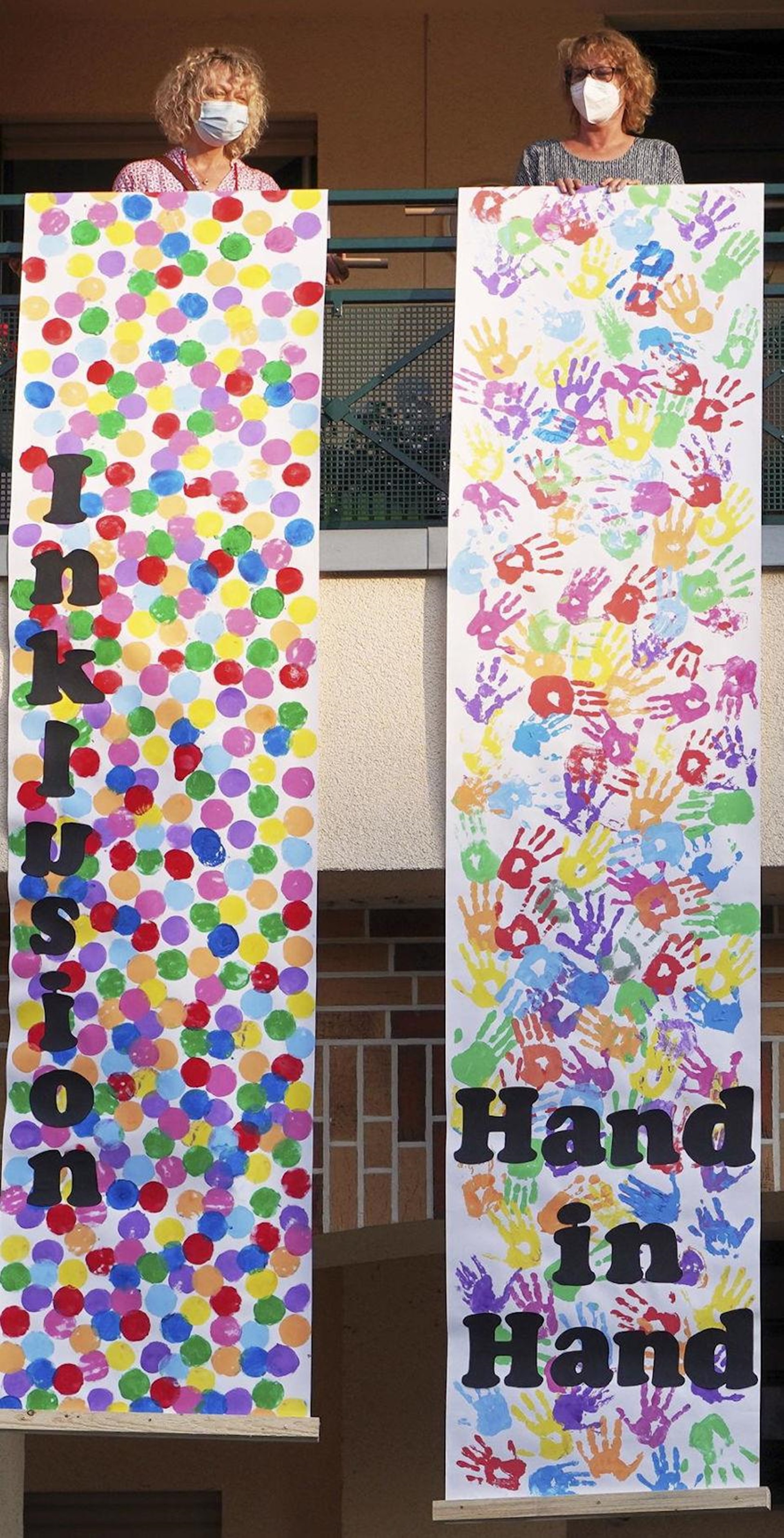 Zwei Frauen stehen auf einem Balkon und lassen bemalte Transparente vom Haus herab.